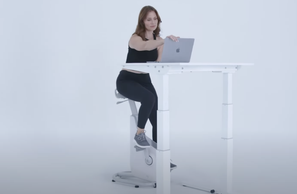 A person in workout clothes uses a stationary bike while working on a laptop at a standing desk. The setting is minimal with a white background.