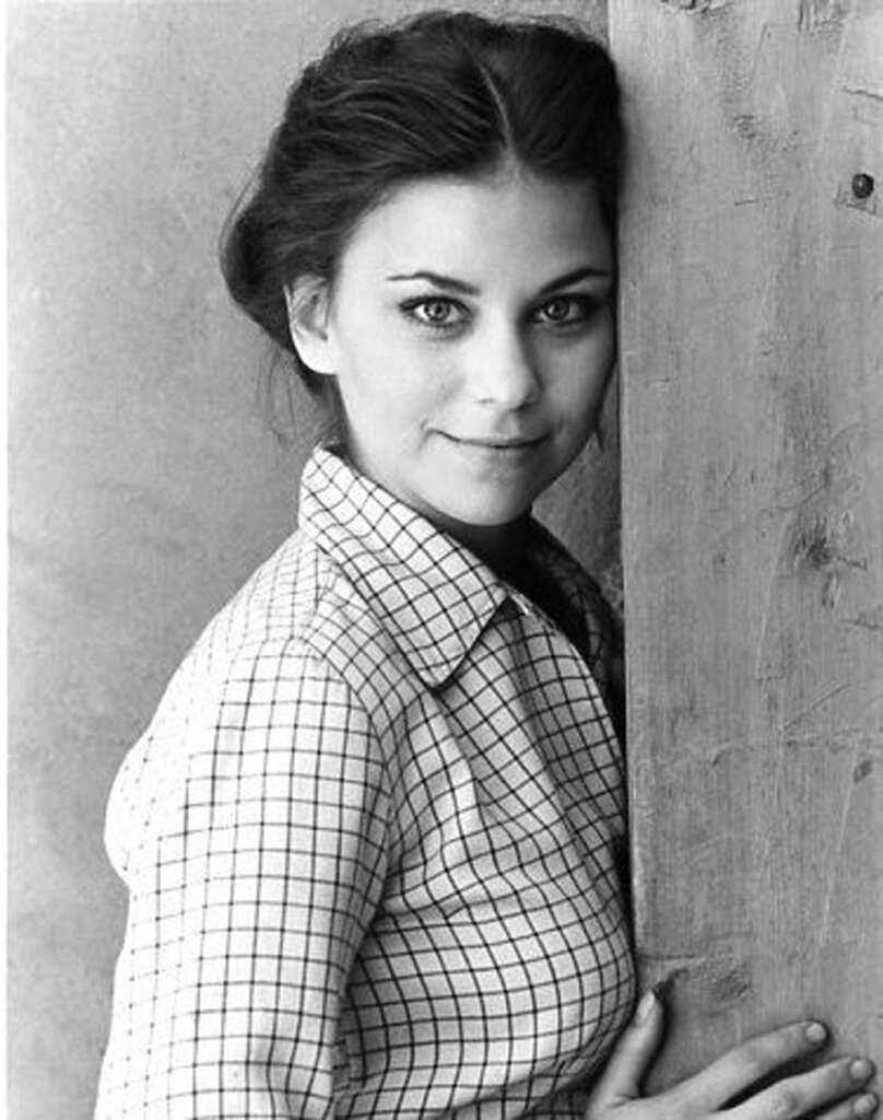 Black and white portrait of a woman with dark hair tied back, wearing a checkered shirt. She is leaning against a wooden surface, looking directly at the camera with a slight smile.