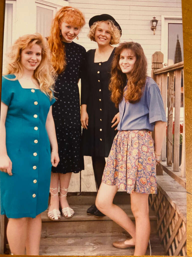 Four women are posing on wooden steps in front of a house. They are smiling and dressed in vintage-style clothing: dresses with buttons, a polka-dot dress, white sandals, and a black hat.