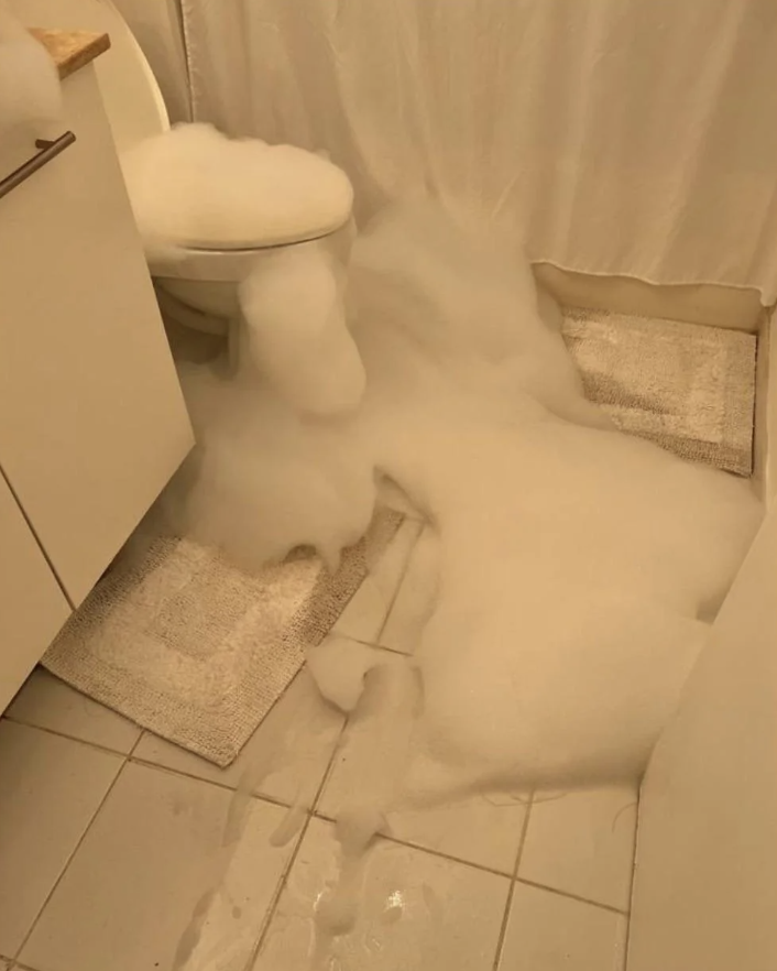 A bathroom scene shows white foam overflowing from the toilet onto the floor, spreading across the tiles and rugs. The foam appears soft and fluffy, creating a dramatic and unusual effect in the small space.