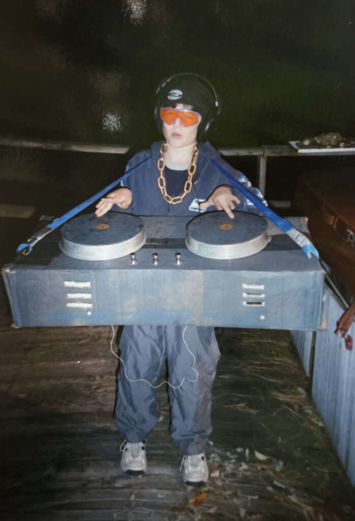 A child dressed as a DJ, wearing orange sunglasses, headphones, and a gold chain, holds a cardboard DJ setup with painted turntables. They are outdoors on a wooden surface, wearing a blue outfit and sneakers.