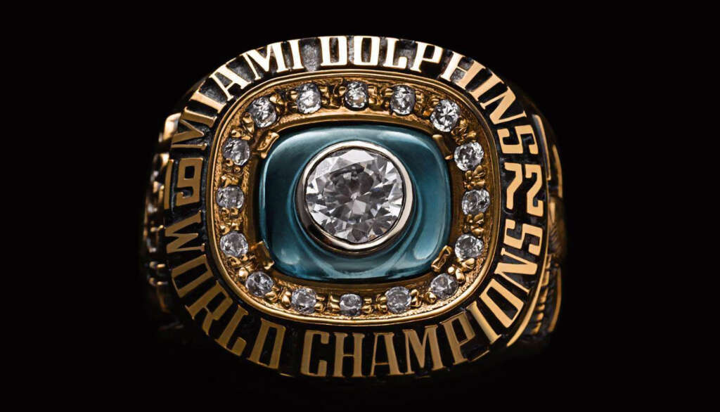 A gold championship ring with a large central diamond, surrounded by smaller diamonds, featuring the text "Miami Dolphins" and "World Champions" engraved around it. A teal background highlights the center stone.