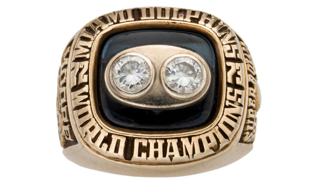 A gold ring with the words "World Champions" engraved around a black center, featuring two large, round diamonds. Engravings are also present on the sides of the ring.