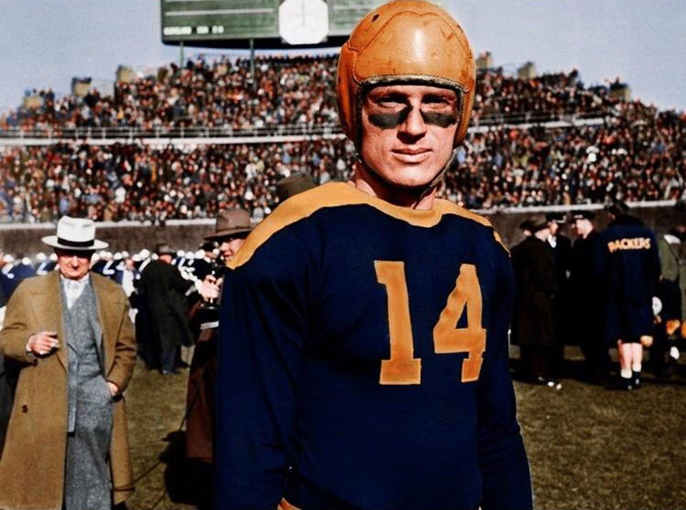 Football player in a vintage uniform and leather helmet stands on a field. He wears a navy jersey with the number 14 and gold accents. A crowd in a stadium and other players are in the background.