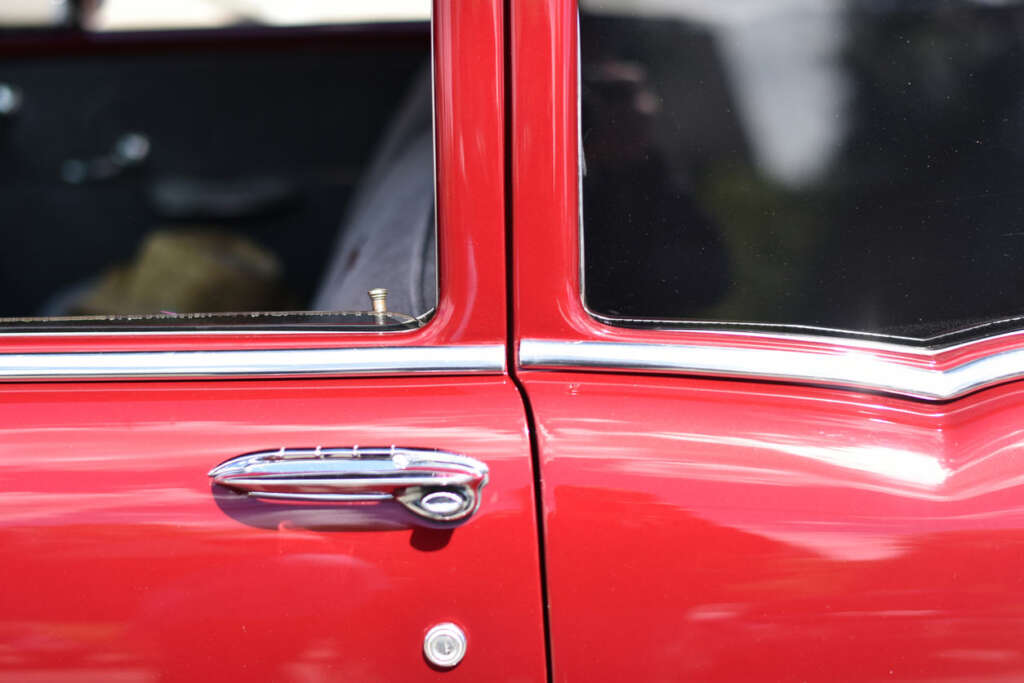 Close-up of a red classic car door highlighting the chrome handle and trim. The window is partially visible, reflecting some outdoor surroundings and sky. The car's glossy paint and elegant design details are showcased.