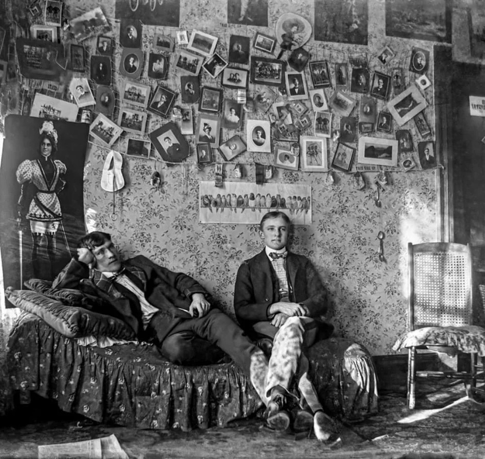 Two men in vintage attire sit in a room with ornate wallpaper. One reclines on a bed, while the other sits on a chair. The walls are covered with numerous photographs and portraits. A woven chair is to the side. The setting exudes a historical ambiance.