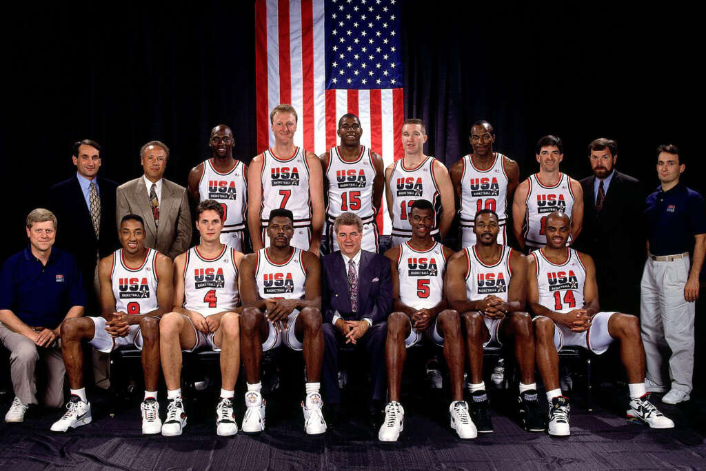 A group photo of a basketball team in USA-themed uniforms. The team members are posing in two rows with coaches. There's an American flag in the background. Some team members are seated, while others stand behind them.