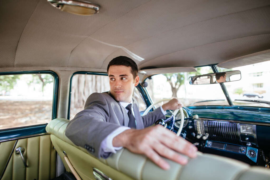 A man in a suit is sitting in the driver's seat of a vintage car, looking over his right shoulder. The car's interior is beige and teal, with classic details. Trees are visible through the windows.