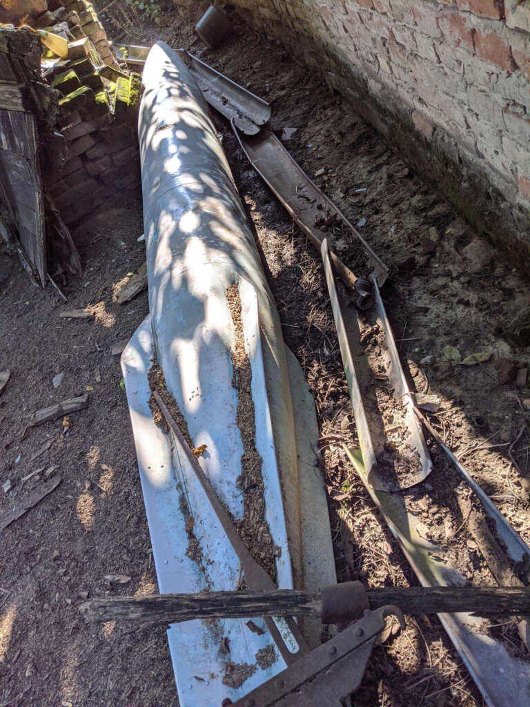 A weathered, long, narrow metal structure lies on the ground, partially covered in dirt. It's surrounded by debris and wooden planks, next to a brick wall. Sunlight casts shadows of tree branches across it.