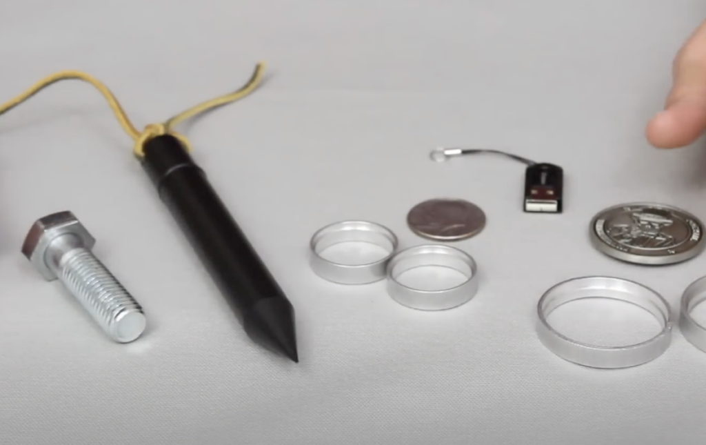 A collection of objects on a surface: a large bolt, a black pen with a yellow cord, several metal rings, a small USB flash drive, a quarter, and a large silver coin. A finger is partially visible on the right side.