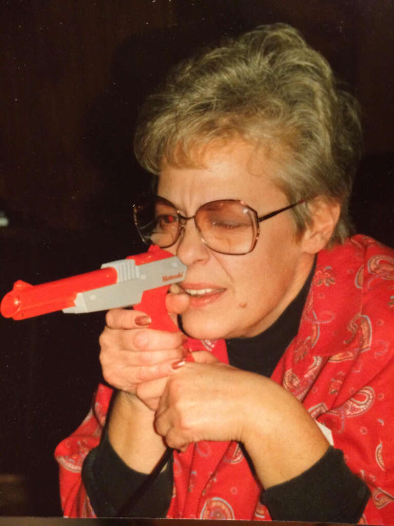 A person with short, light hair and glasses is aiming a Nintendo Zapper gun, part of the Nintendo Entertainment System, while wearing a red patterned jacket. The person is focused, with one eye closed.