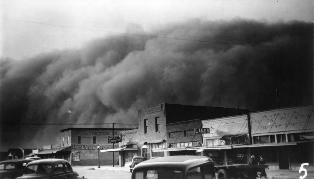 Black and white photo of a looming dust storm approaching a small town street with brick and wooden buildings. Vintage cars are parked along the road, and the sky is dark with approaching clouds.