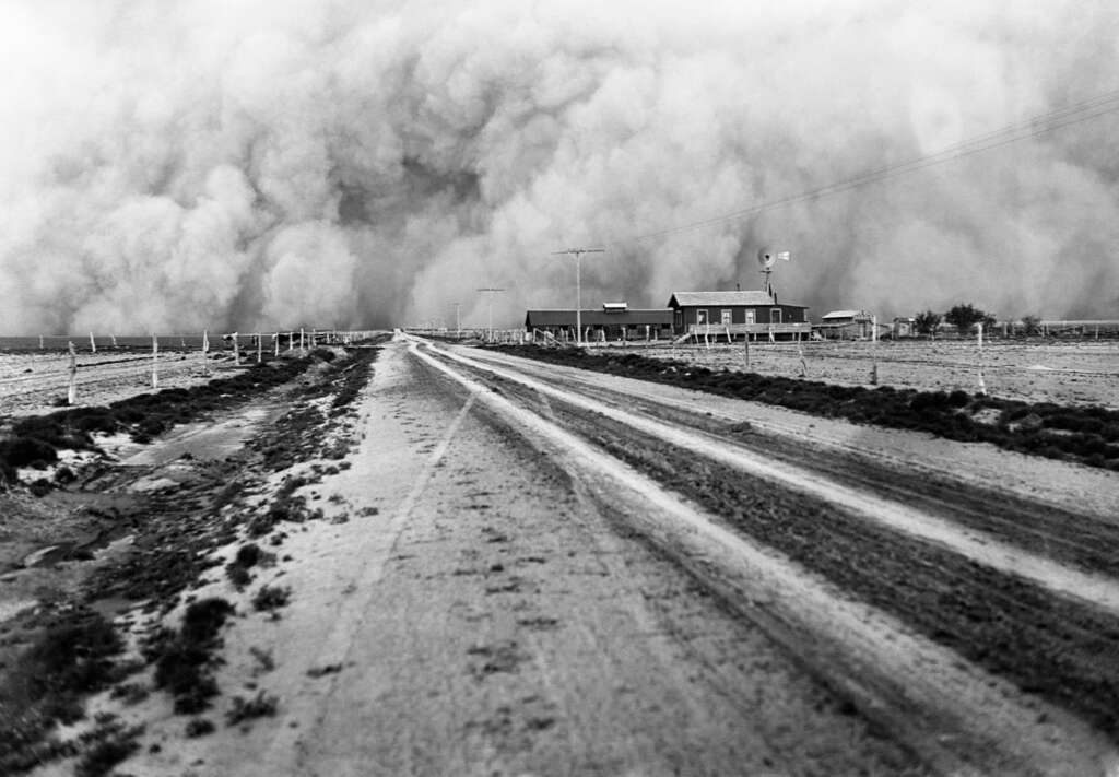 A black and white photo shows a vast, flat landscape with a long, dirt road leading to a small farmhouse. A massive, thick dust storm looms in the background, engulfing the sky and approaching the house. Power lines stretch along the road.