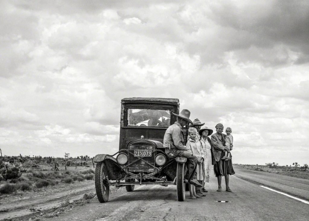 Black and white photo of a family beside an old car on a deserted road. A man in a hat sits on the car's running board, while a woman holds a child. Two other women stand nearby, all gazing off into the distance under a cloudy sky.