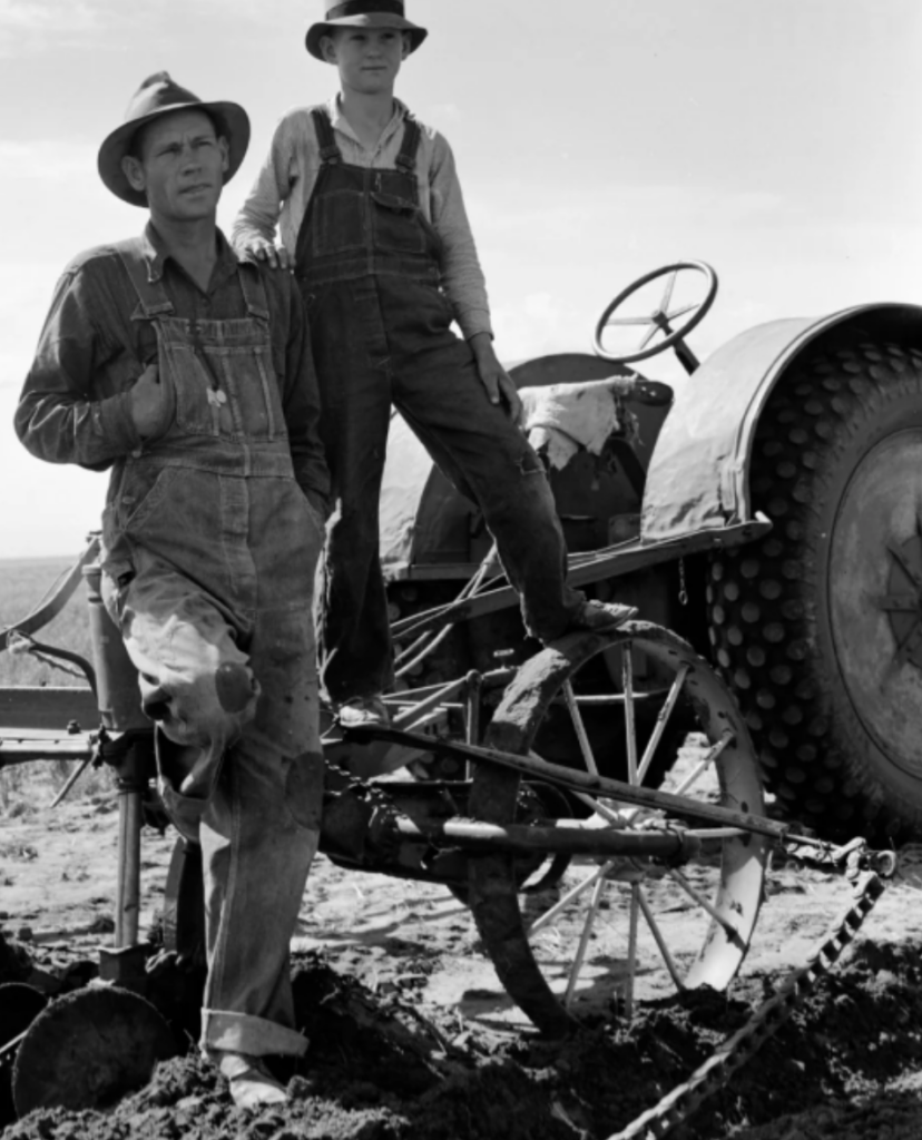 Two men in overalls and wide-brimmed hats stand beside a farming tractor on a field. One man leans against a large wheel while the other sits on the tractor. The background shows a clear sky and plowed soil. The scene is in black and white.