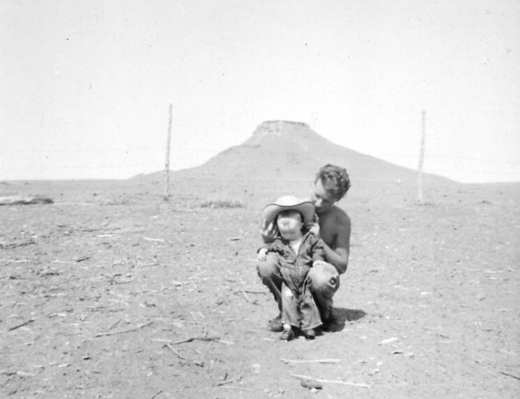 A man crouches behind a young child dressed in a cowboy hat and boots, in an open, barren landscape. A mesa is visible in the background, with a wire fence cutting across the scene. Both are looking downward.