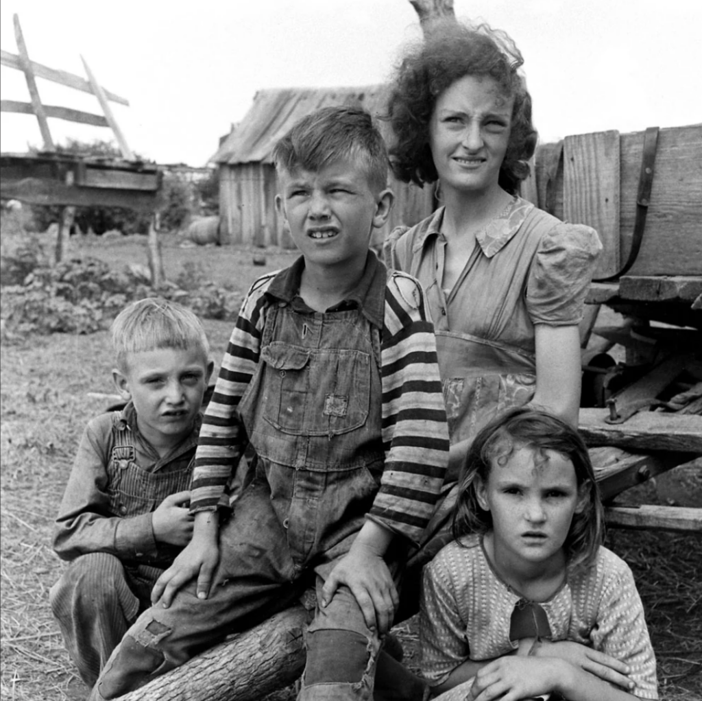 A black and white photo shows a woman with three children in a rural setting. The two boys and one girl sit or kneel on the ground in front of her. A wooden fence and building are visible in the background. All are wearing simple, worn clothing.