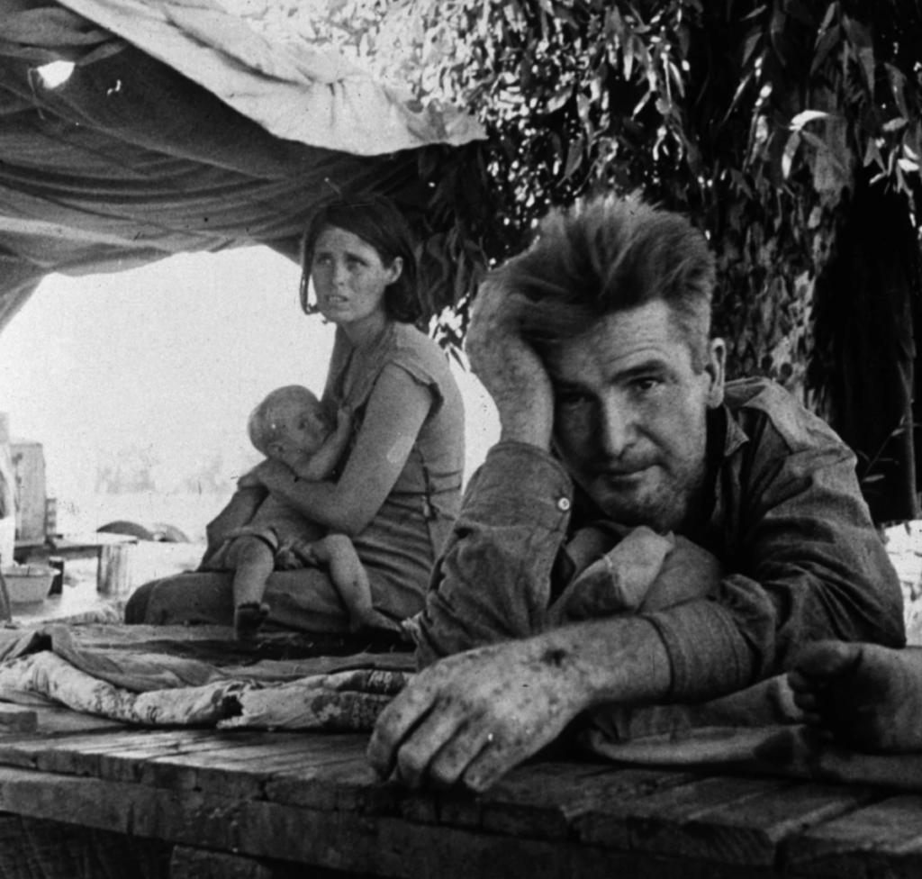 A tired man leans on a wooden table, resting his head on his hand. Behind him, a woman holds a child on her lap under a makeshift tent. Sunlight filters through, highlighting their worn, rugged clothing and the weary expressions on their faces.