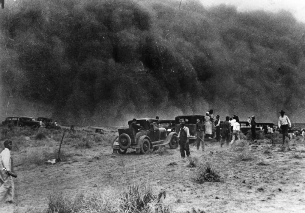 A historical black and white photo shows people and cars fleeing from an approaching dust storm. The ominous cloud looms large in the background, while individuals hurry on dusty ground in the foreground. The scene conveys urgency and tension.