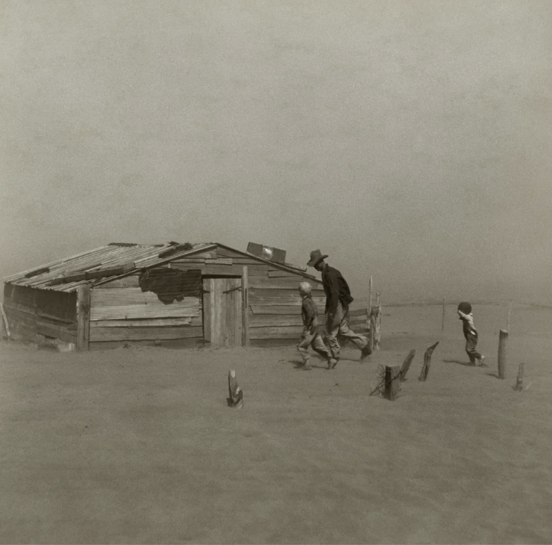 A man with two children walks in a barren, dusty landscape toward a weathered wooden shack. The ground is covered in sand or dirt, and the sky appears hazy, suggesting harsh environmental conditions.