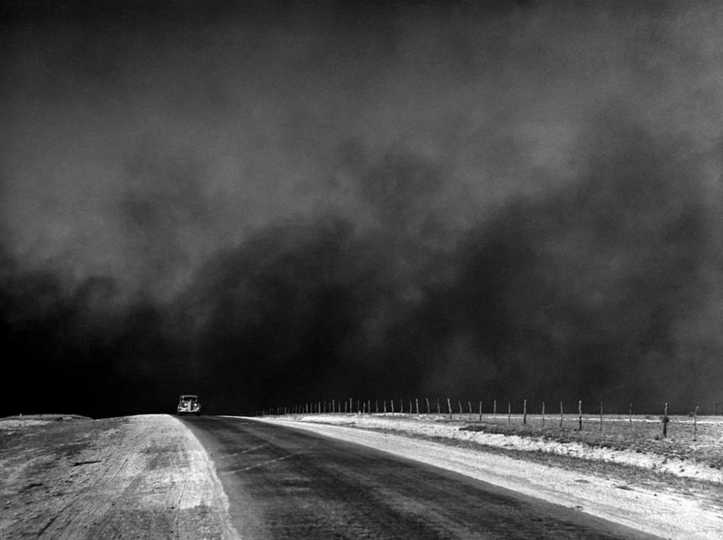 A vintage car drives on a deserted road under a dark, turbulent sky filled with dust and clouds. The landscape is barren, bordered by a wire fence. The scene captures an intense and foreboding atmosphere.