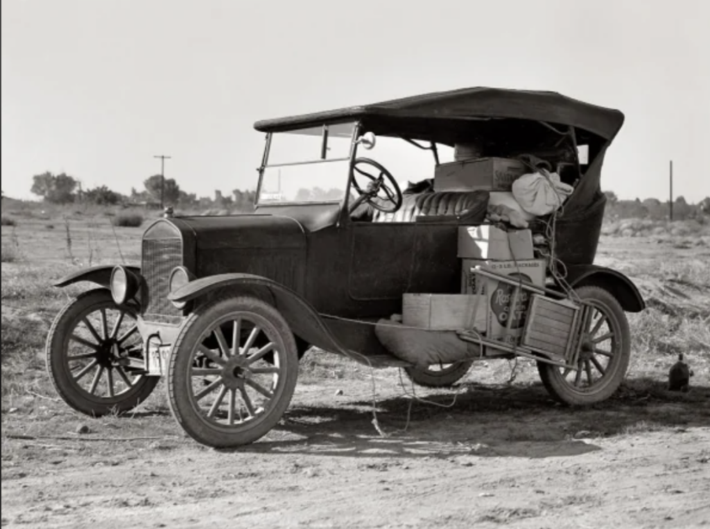 A vintage car with a soft top is parked on a dirt road, laden with various boxes and bags strapped to its sides and back. The landscape is open and barren, with distant trees and telephone poles visible.