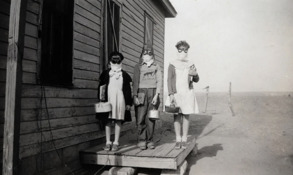 Three children stand on a wooden platform next to a house, wearing goggles and masks, holding lunch boxes. The setting appears dusty and rural, suggesting a scene from the Dust Bowl era.