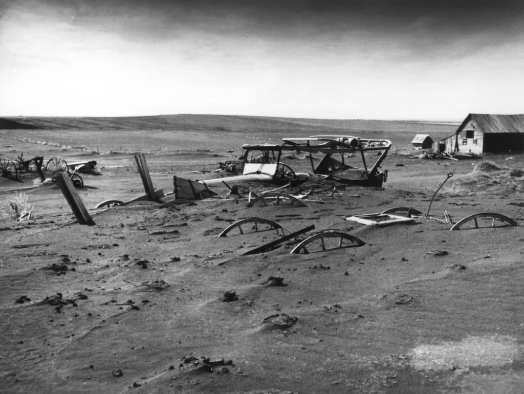 A black-and-white photo shows a barren landscape during the Dust Bowl. Abandoned, partially buried cars and wooden debris are scattered in the dirt. A few small buildings are visible in the background under a cloudy sky.