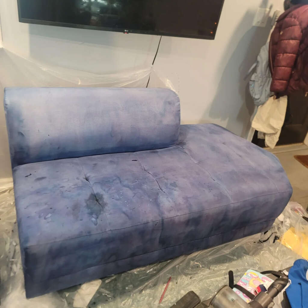 A blue upholstered chaise lounge with a backrest against a white wall. A TV is mounted above it. Plastic covers the floor, with items like tools nearby. Jackets hang on hooks on the wall to the right. The chaise has some visible stains.