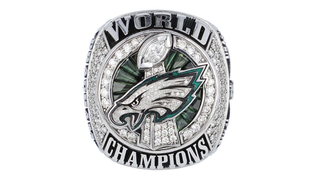 A championship ring featuring a prominent eagle emblem at the center, surrounded by green gemstones. The words "WORLD CHAMPIONS" are engraved along the top and bottom in bold letters. The ring is adorned with numerous sparkling diamonds.