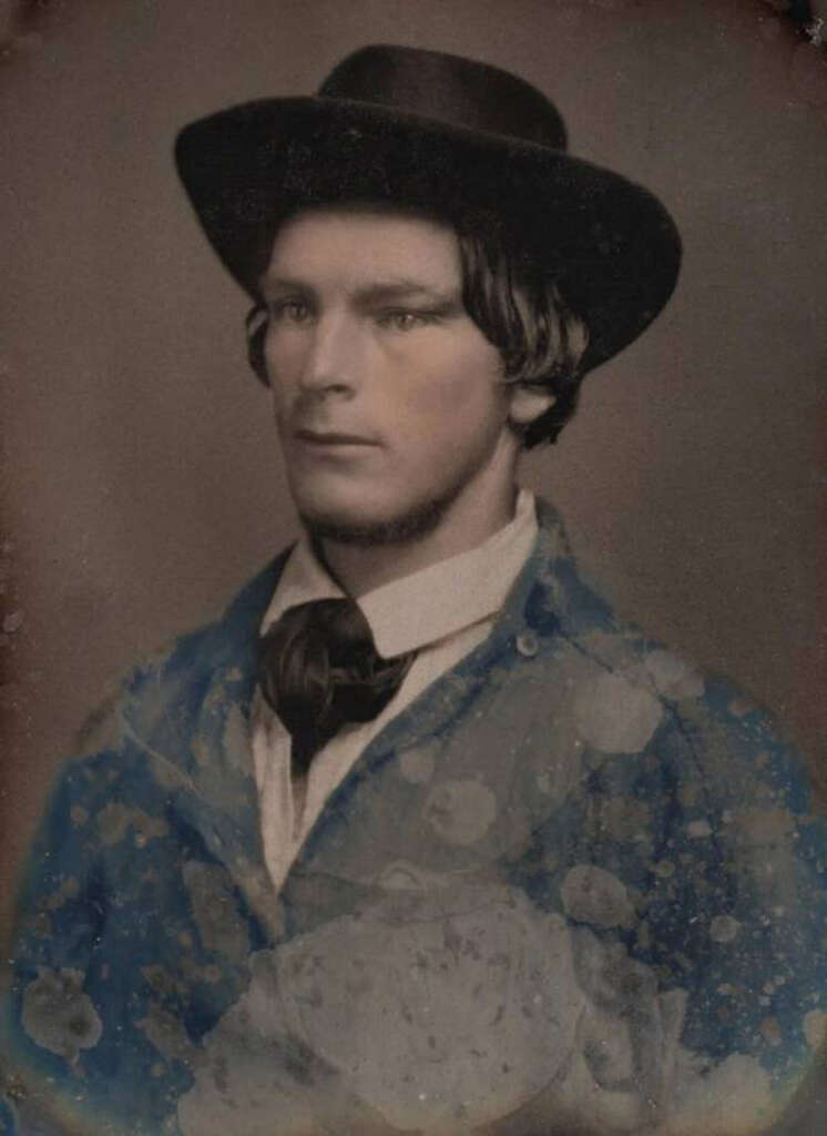 A sepia-toned vintage portrait of a man wearing a wide-brimmed hat and a tie. He has dark hair and is dressed in a formal jacket. The image shows signs of aging, with visible spots and discoloration.