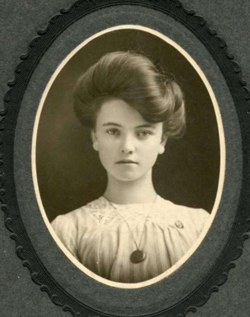 Vintage oval-framed portrait of a young woman with styled hair wearing a high-collared blouse and a pendant necklace. She gazes directly at the camera with a neutral expression. The background is plain, typical of early 20th-century photographs.