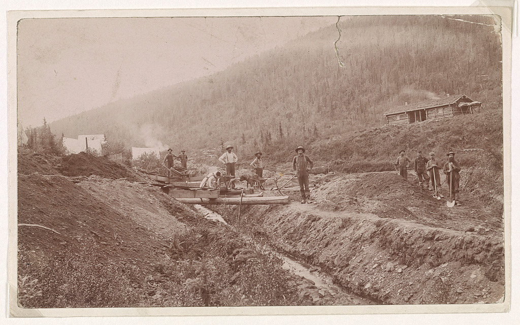 A vintage photo of eight men working on a hillside excavation with shovels and wheelbarrows, near a wooden cabin and a stream. Trees cover the hill in the background. The scene appears to be from a historical mining or construction site.