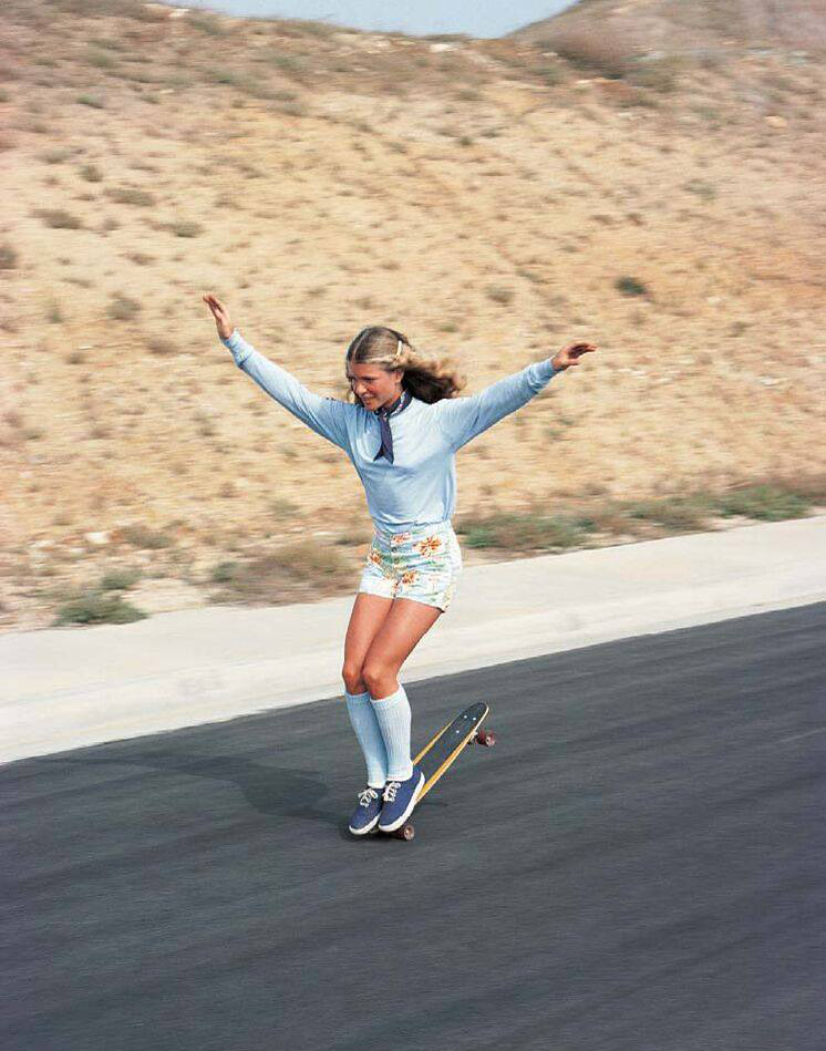 A person skateboards down a paved road with arms outstretched for balance. They wear a light blue long-sleeve shirt, floral shorts, knee-high socks, and sneakers. The background features dry grass and hills.