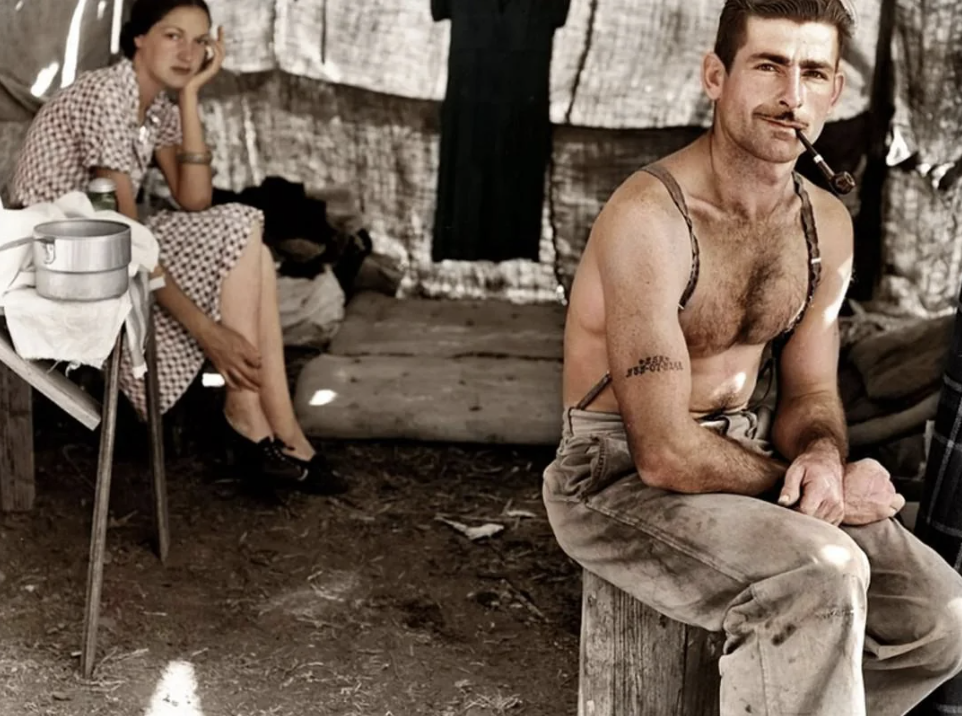 A man with a pipe, wearing suspenders and work pants, sits on a wooden block in front of a makeshift tent. A woman in a checkered dress sits behind him, resting her chin on her hand. A pot and other items are on a nearby table.