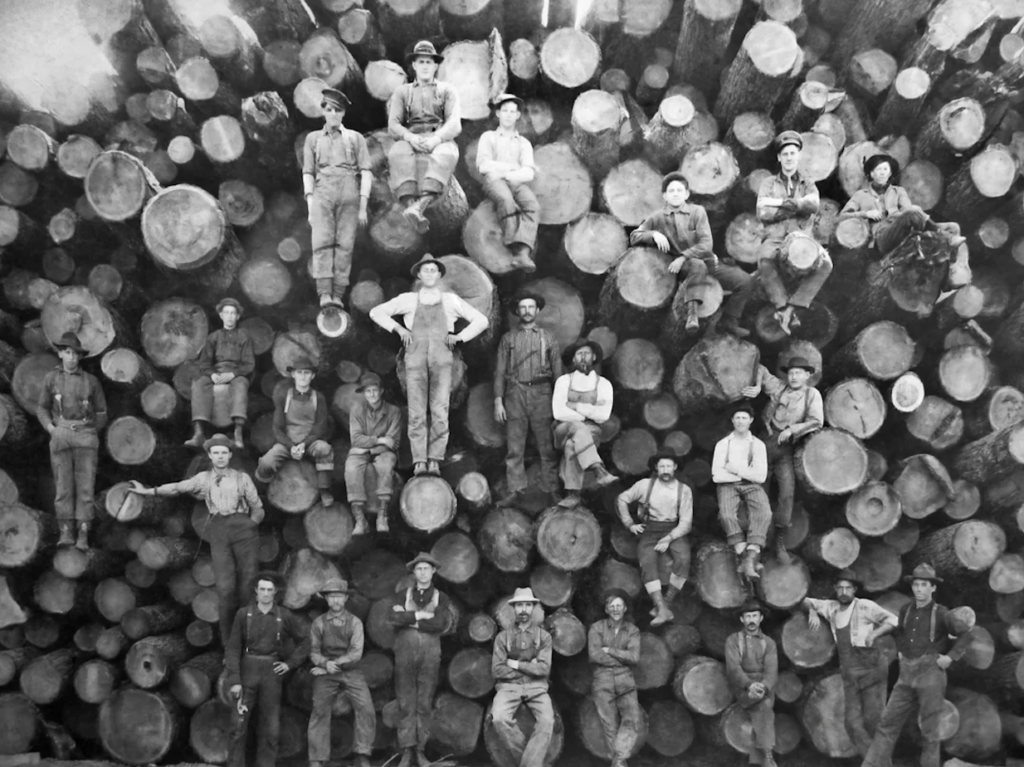 A group of men in work attire, posing on a large stack of logs. They are arranged at different heights, showcasing the massive size of the logs. The image is in black and white, suggesting it is from an earlier time period.