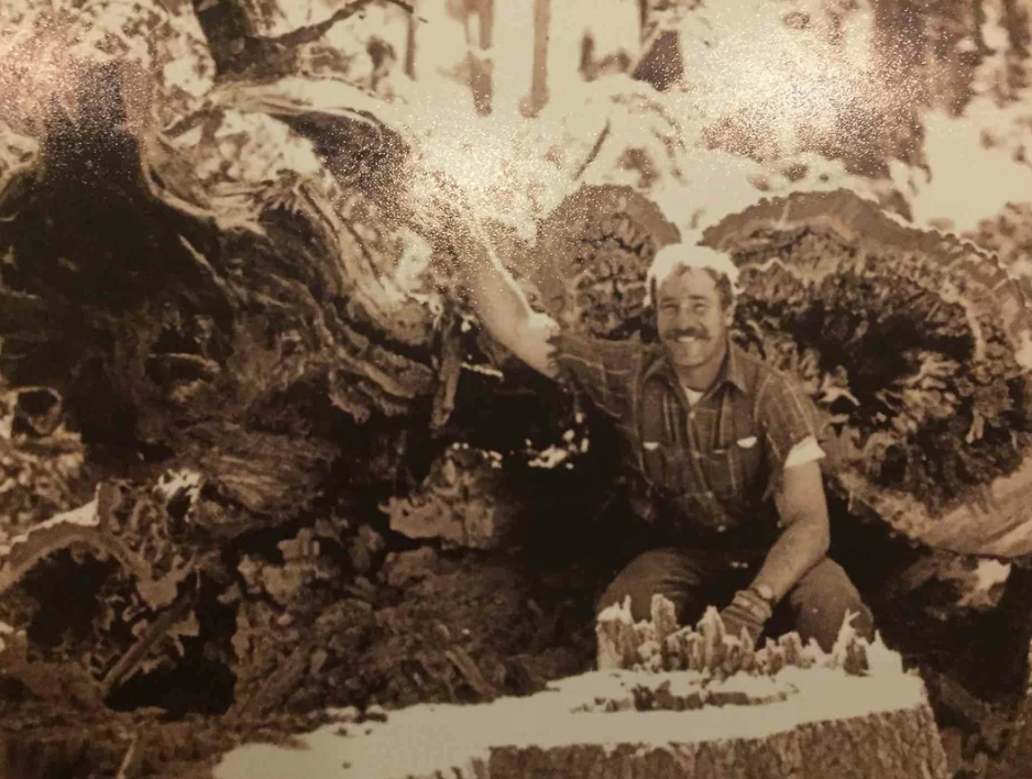 A man with a mustache in a plaid shirt is kneeling beside a large, fallen tree trunk, smiling and holding onto it. The background shows a forest setting with more trees. The image is in sepia tones.