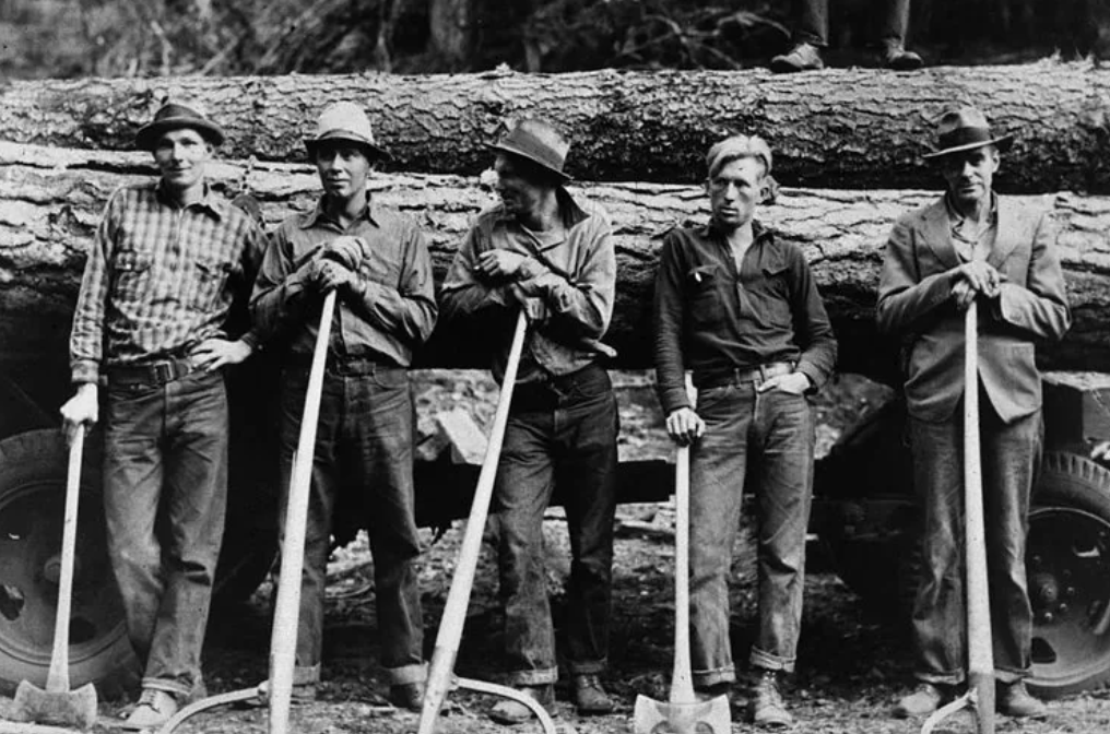 Five lumberjacks stand in front of a large log resting on a truck, each holding an axe. They wear hats and work clothes, looking directly at the camera. Trees and foliage are visible in the background.