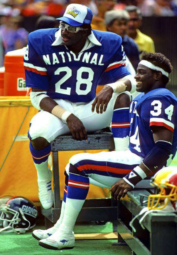 Two American football players in blue jerseys with "NATIONAL" and numbers 28 and 34 sit on the sidelines. They are wearing helmets and are seated on a bench during a game. One player sports a visor cap, the other a headband.