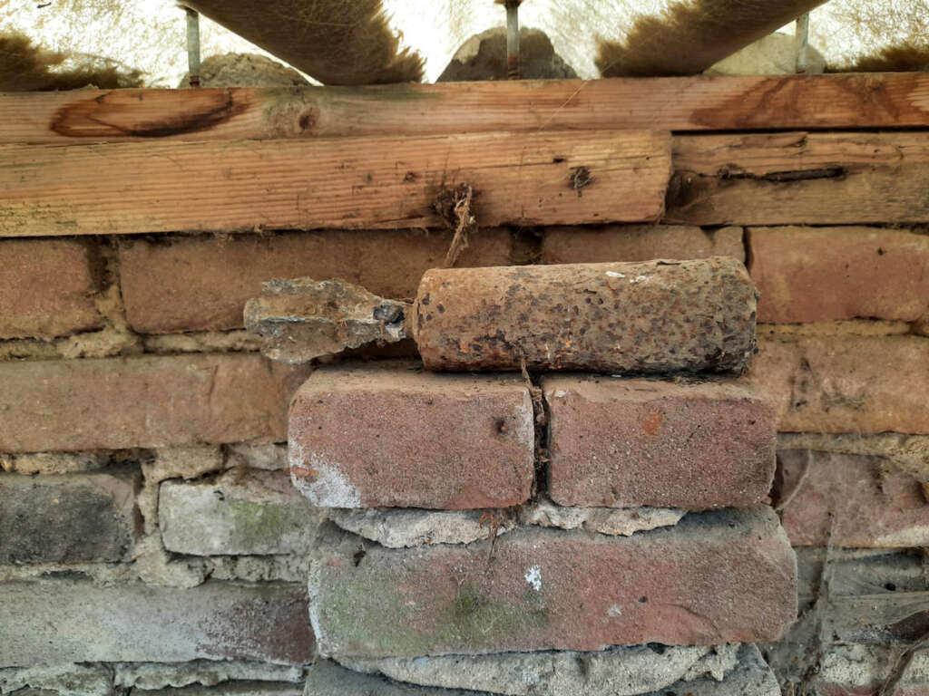 A rusted, cylindrical object is resting on a rough brick wall. The wall is topped with a wooden beam, and the area appears weathered and aged. The background includes a partially visible roof structure.