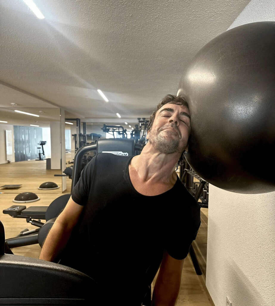 A man wearing a black shirt leans against a large exercise ball in a gym. The gym has wooden floors and several exercise machines in the background. The man appears relaxed with his eyes closed.