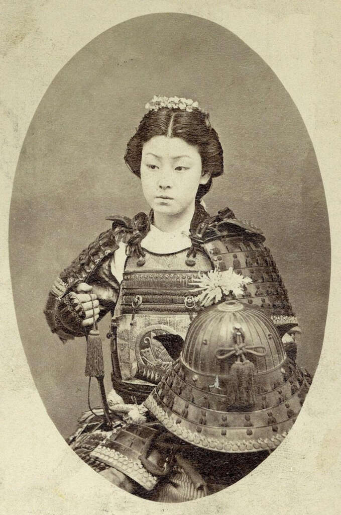 Black and white photo of a young woman wearing traditional samurai armor, holding a samurai helmet. Her hair is styled with floral adornments. The background is plain and oval-shaped, highlighting her poised expression and historical attire.