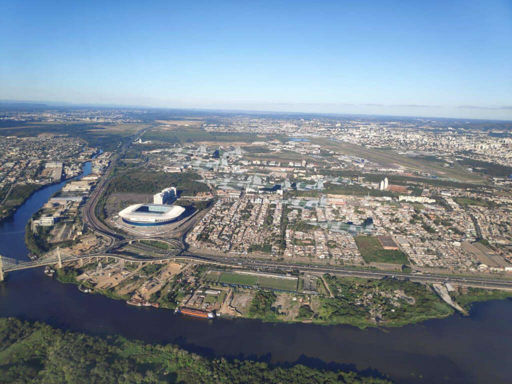 Aerial view of a sprawling cityscape with a large stadium, river, and expansive residential and industrial areas. Roads and bridges intersect, surrounded by greenery under a clear blue sky.