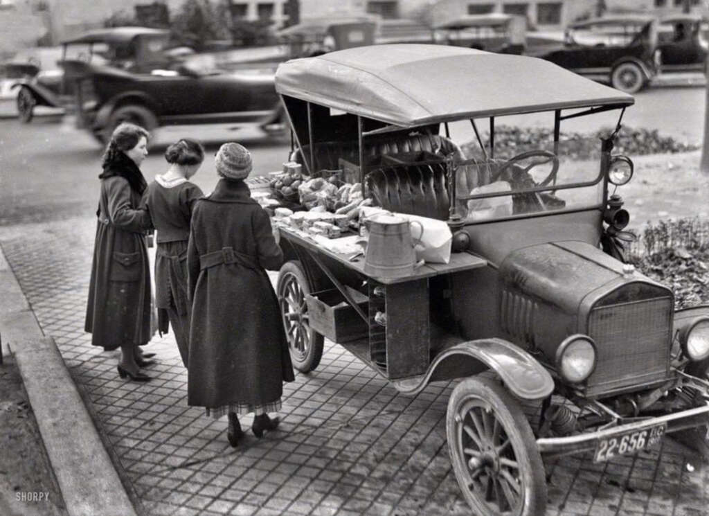 Three women browse a variety of goods displayed on the back of a vintage truck parked on a brick sidewalk. The truck is filled with items like fruits, vegetables, and household goods. Another car is blurred in motion in the background.