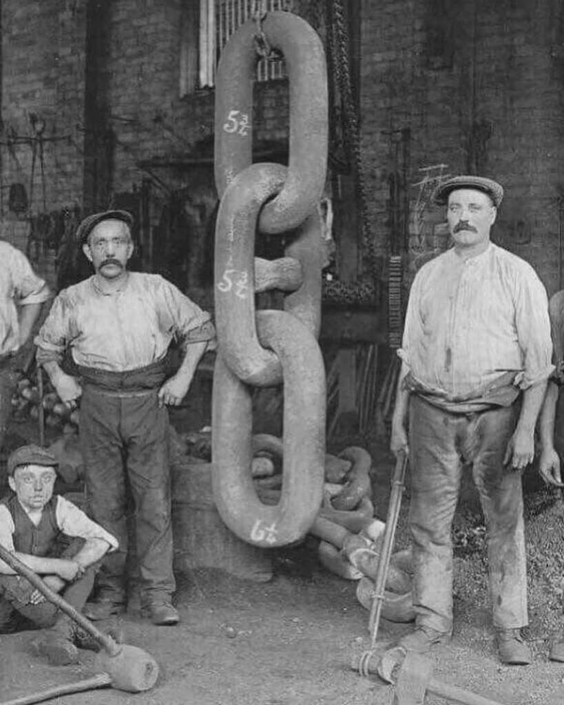 Two men in early 20th-century attire stand beside a large chain link in a workshop. Both wear flat caps and shirts, holding tools. An additional man crouches nearby. The chain links have visible measurements.