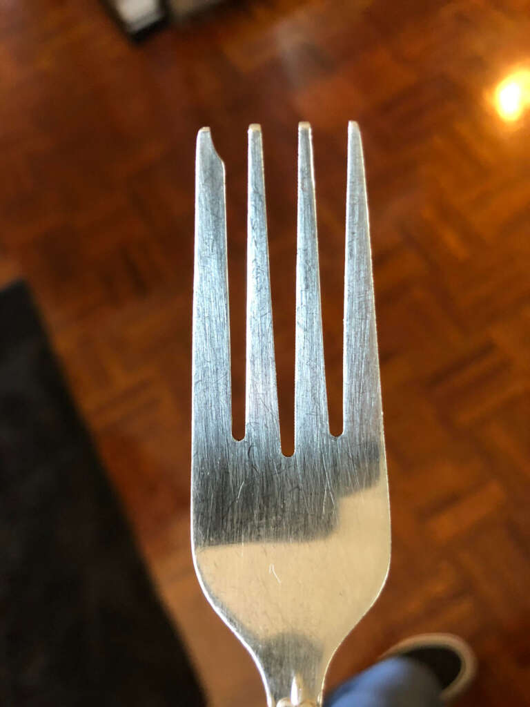 Close-up of a silver fork with four tines, where the leftmost tine is chipped at the tip. The wooden floor in the background has a warm tone.