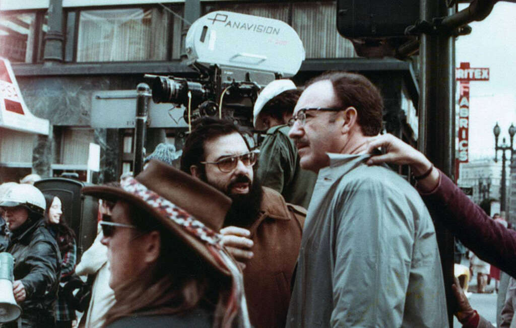 A group of people stands on a bustling city street. A man with glasses and a mustache speaks animatedly to another man in glasses and a coat. A large Panavision camera is set up in the background for filming. Passersby and crew members are visible.