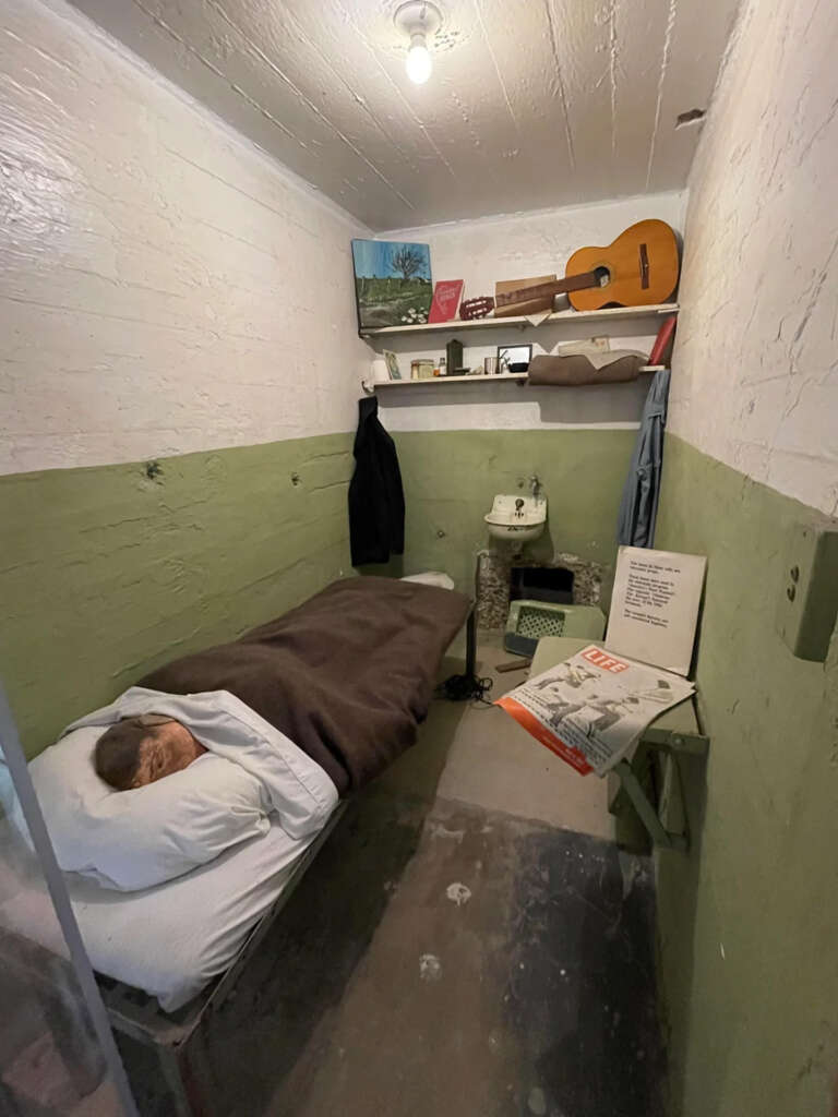 A narrow prison cell with green and white walls, containing a bed with a dummy under a brown blanket. The cell has a shelf with books, a guitar, and art supplies. A jacket hangs on the wall, and there is a small sink and toilet in the corner.