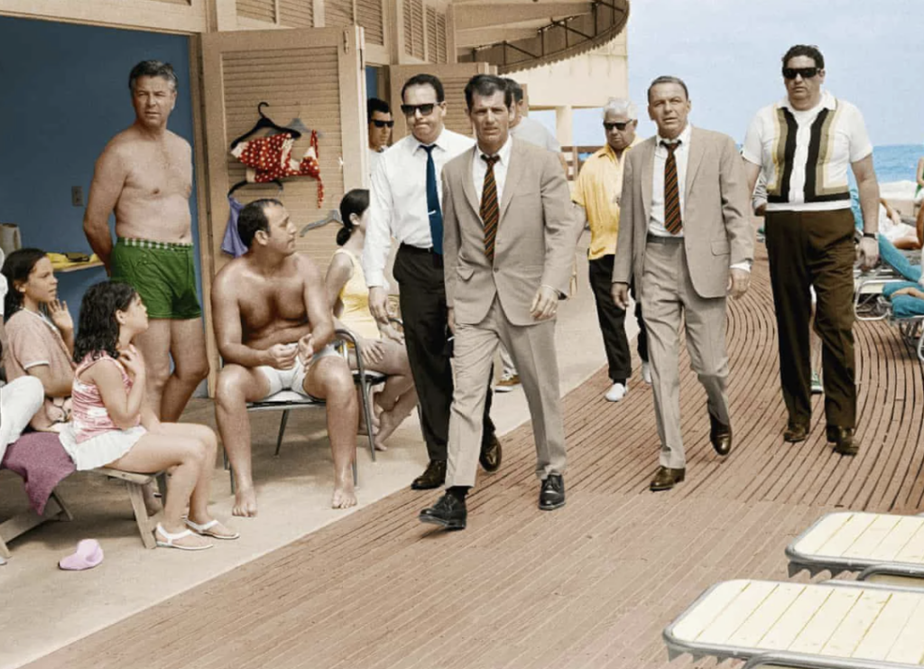 A group of men in suits walks along a wooden deck beside a beach. They are accompanied by others in casual attire, including a man in green swim shorts seated with women. The ocean is visible in the background.