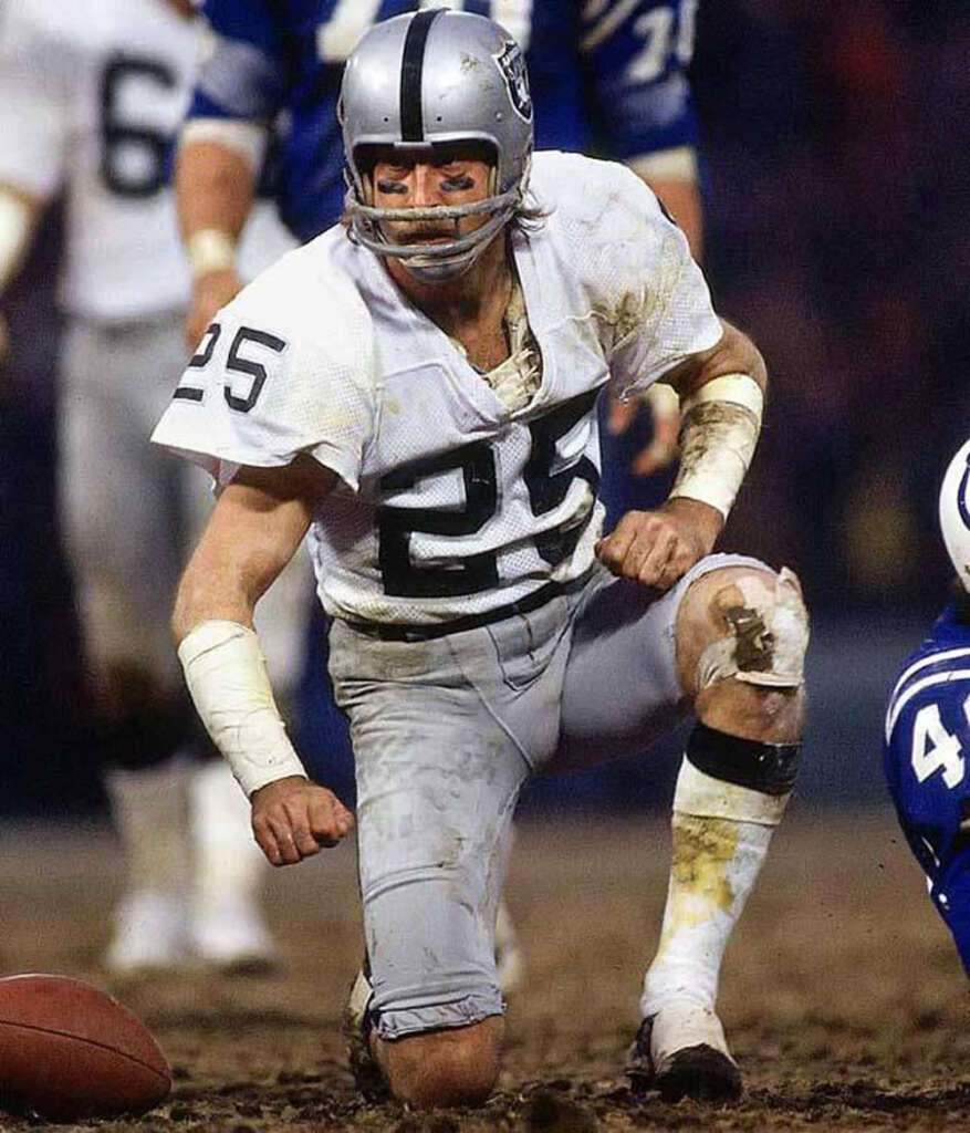 Football player in a white and silver uniform with the number 25 kneels on a muddy field, looking intensely forward. The player wears a silver helmet with a logo on the side. Another player in blue is partially visible.